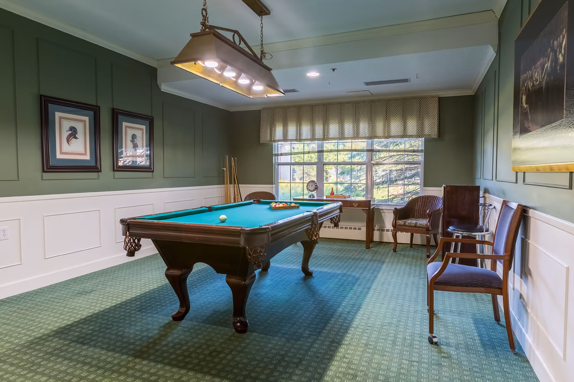 A well-lit room with a green carpet and green walls featuring a pool table in the center. The pool table has balls arranged for a game and cues resting against the wall. There are framed pictures on the wall, a window with a valance letting in natural light, and several chairs arranged around the room.