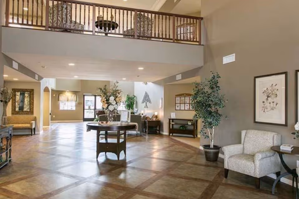 Spacious and well-lit senior living facility common area with a high ceiling and a balcony railing above. The room features a round table with a floral arrangement in the center, comfortable seating including armchairs, framed botanical artwork on the walls, potted plants, and a large window and door letting in natural light.