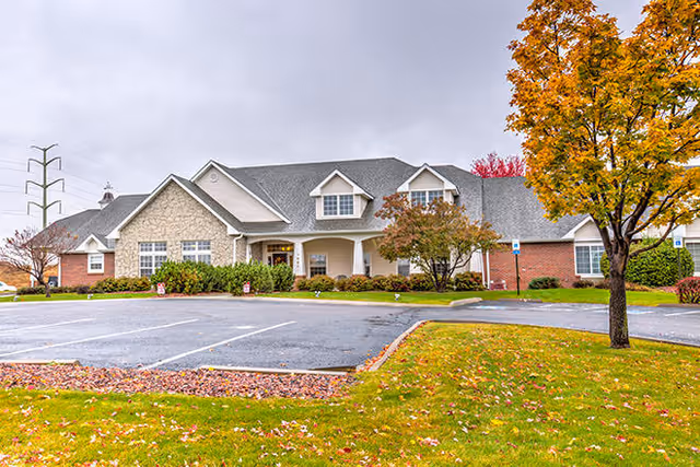 Exterior view of a single-story senior living facility building with a combination of brick and stone facade, a gray shingled roof, and several windows. The foreground shows a parking lot with marked spaces and a grassy area with autumn leaves and a tree with orange foliage. The sky is overcast.