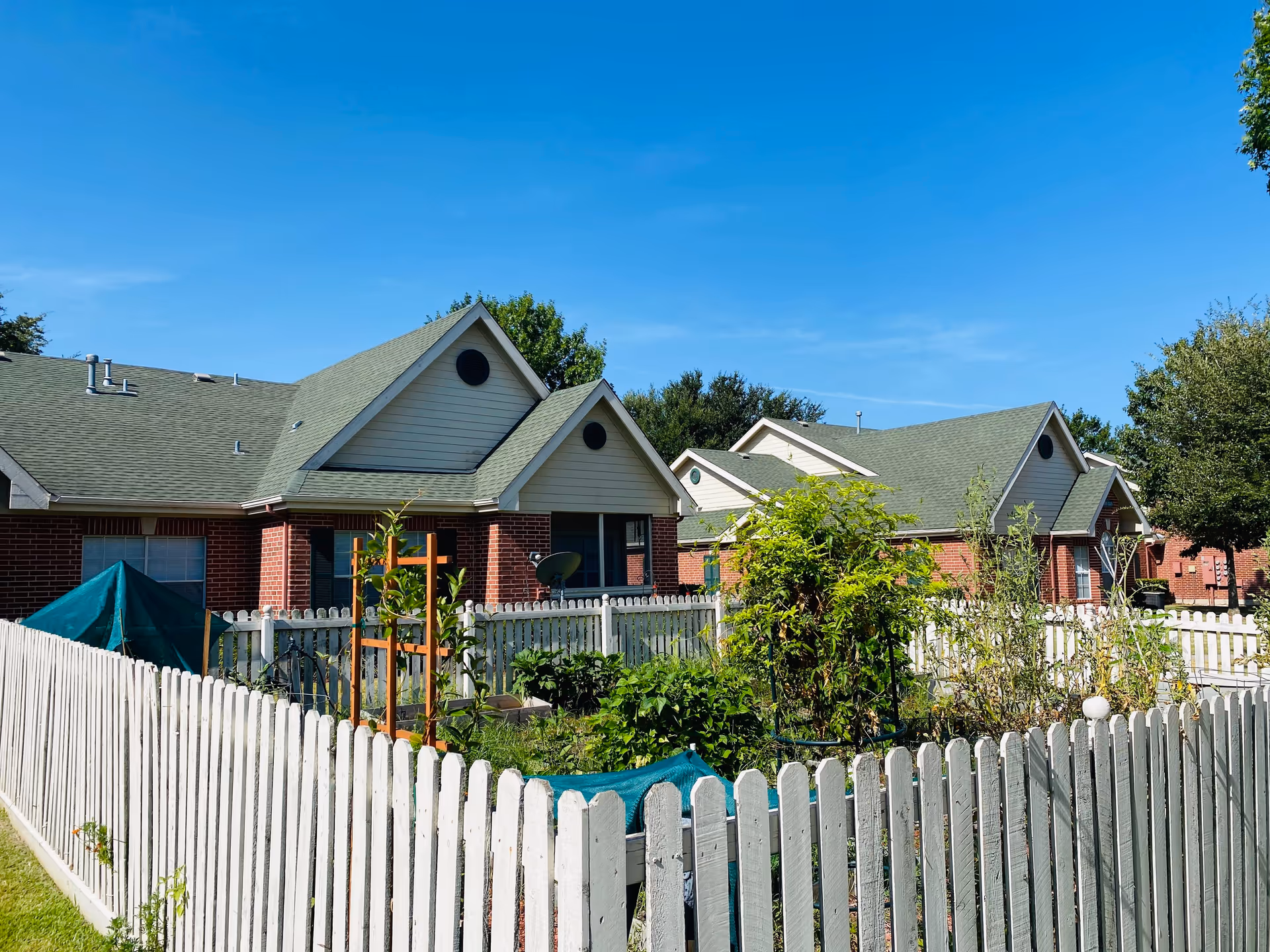 View of a residential area at The Wellington featuring single-story brick buildings with green roofs, surrounded by white picket fences enclosing garden areas with various plants and greenery under a clear blue sky.
