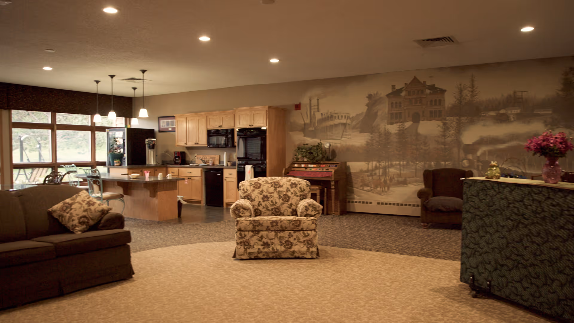 Open common area with sofas and a patterned armchair facing a kitchenette and a large muraled wall.