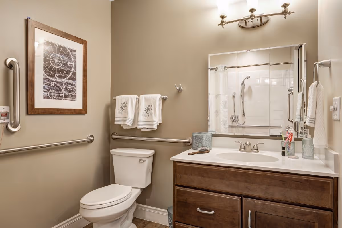 A clean and well-lit bathroom with beige walls, a white toilet, a wooden vanity with a white countertop and sink, a large mirror above the sink, and a shower with a white curtain visible in the reflection. The bathroom has safety grab bars on the walls, two white towels hanging on a towel rack, a framed artwork on the wall, and various toiletries on the countertop.