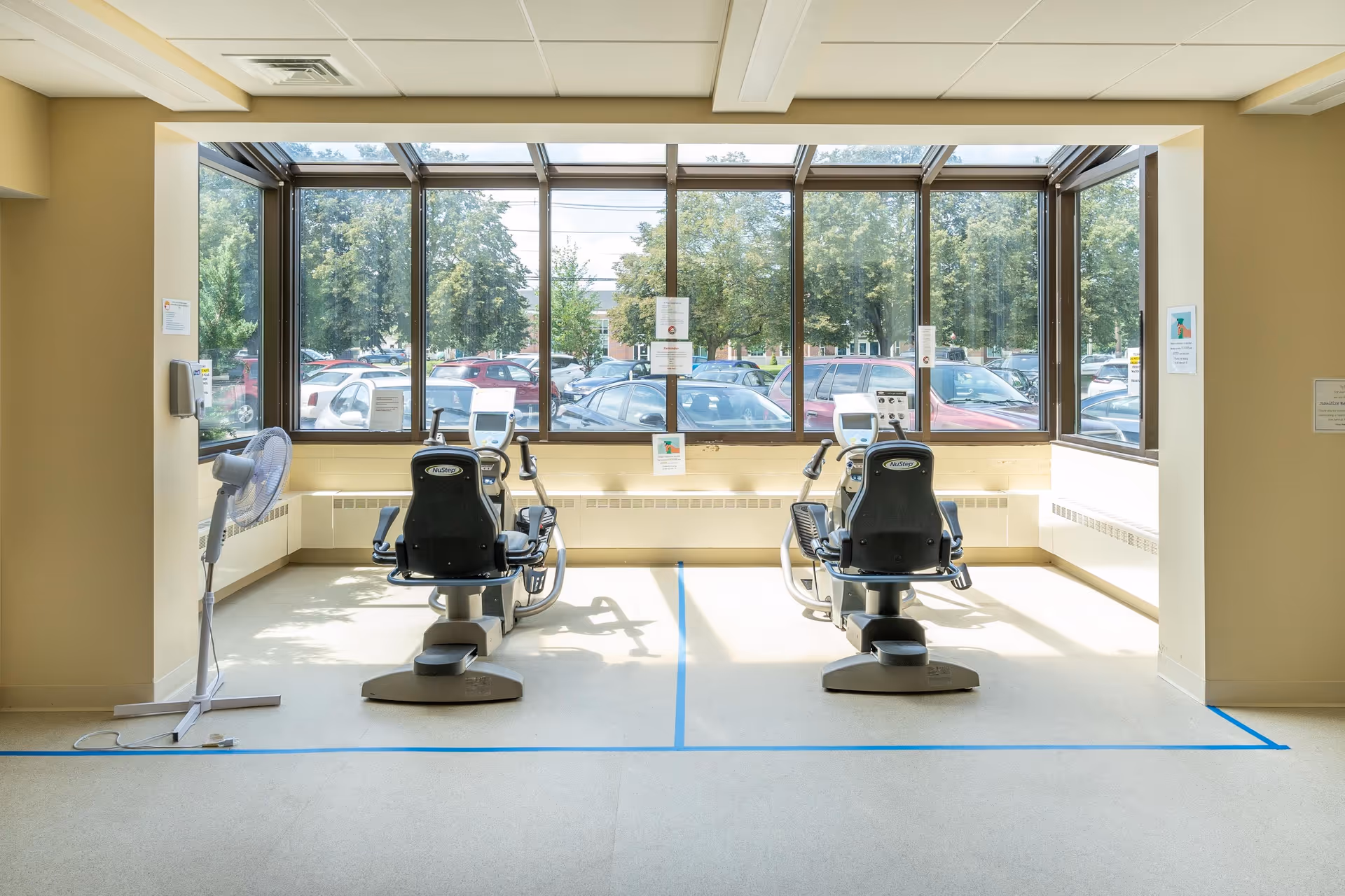 Sunlit exercise area with two recumbent exercise bikes facing large windows overlooking a parking lot.