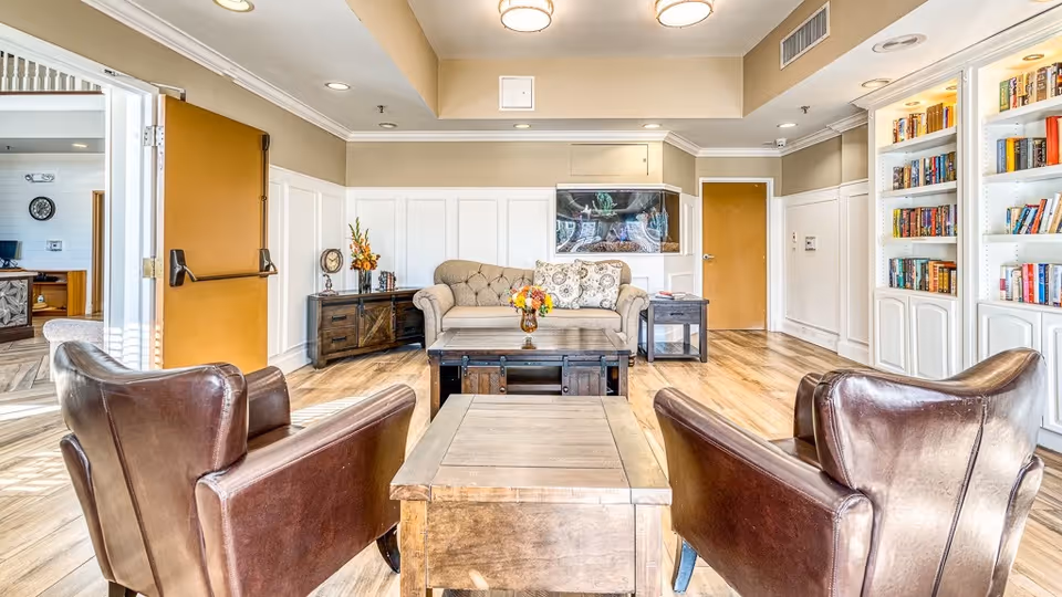 A bright and cozy senior living common area featuring two brown leather armchairs facing a wooden coffee table, a beige tufted sofa with patterned cushions, a side table with a flower vase, a wooden cabinet with decorative items, a built-in bookshelf filled with books, and a large fish tank mounted on the wall. The room has light wood flooring, beige walls with white wainscoting, and recessed ceiling lights.