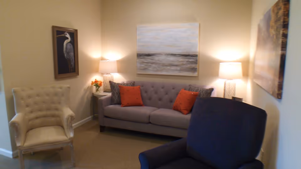 A cozy living room with a gray tufted sofa adorned with two orange pillows and two patterned pillows. There are two table lamps on either side of the sofa, each on a small side table. A beige armchair is on the left and a dark blue armchair is in the foreground on the right. The walls are decorated with three paintings, including one of a bird and two abstract landscapes.