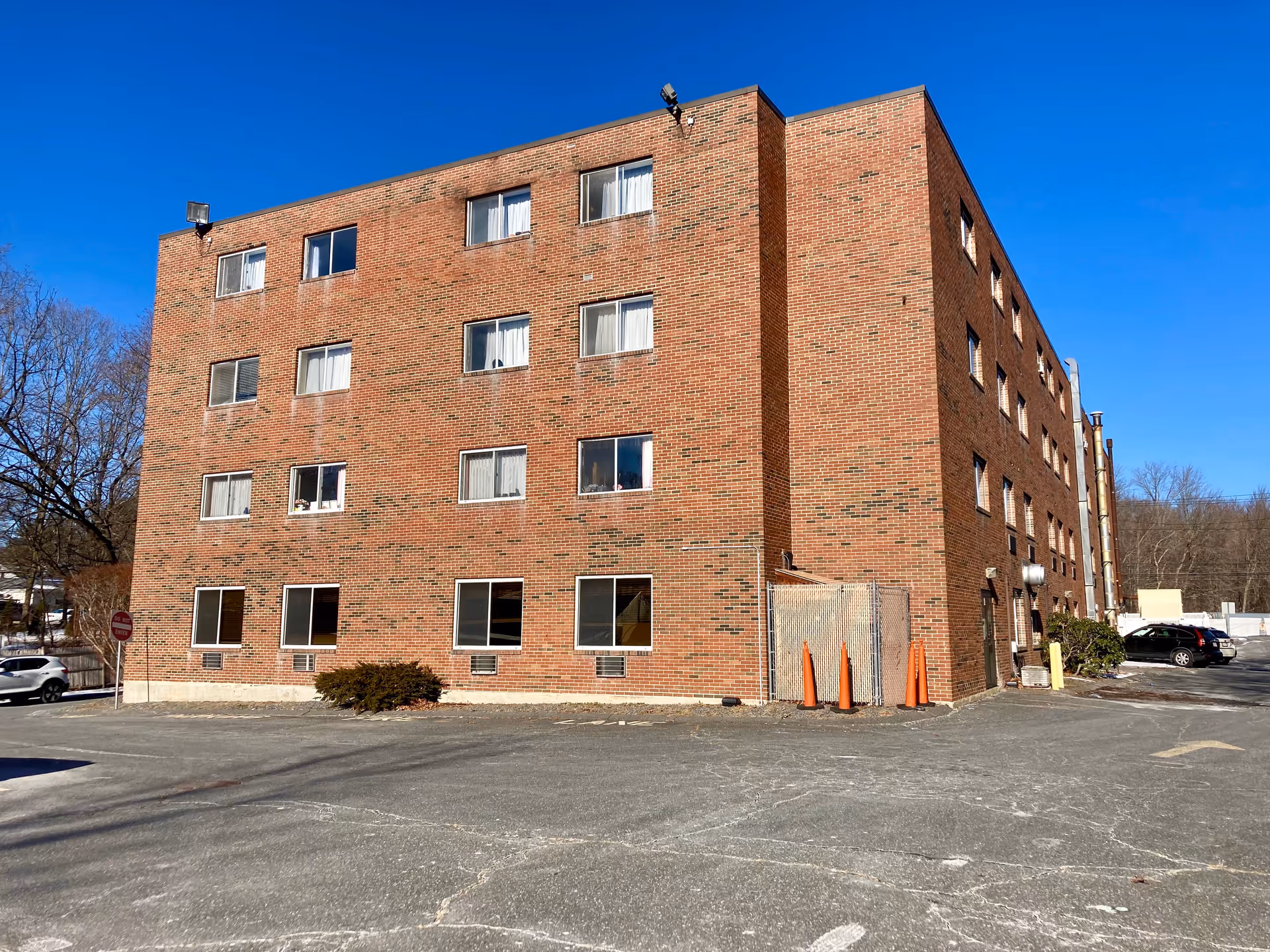 Exterior view of a four-story brick building under a clear blue sky with several windows and a parking area in front. There are a few cars parked and some orange traffic cones near a fenced area on the side of the building.
