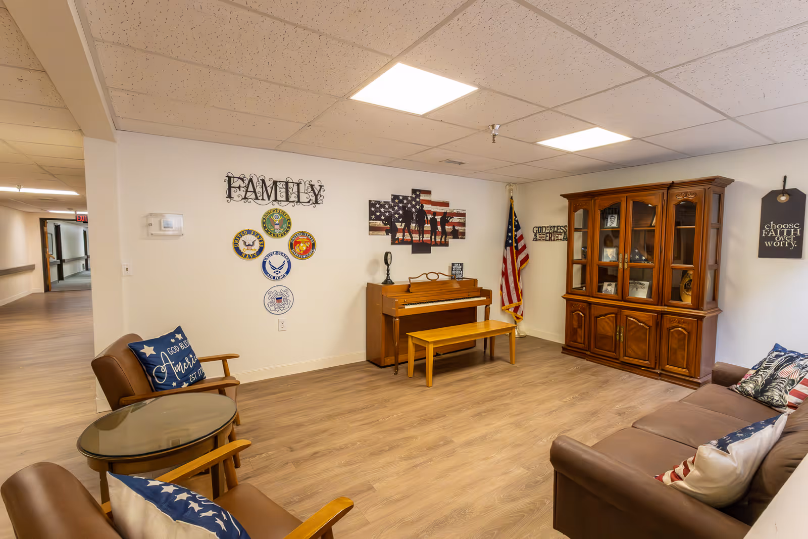 A cozy living room area in a senior living facility with wooden flooring, a brown sofa with patriotic-themed pillows, two brown armchairs with blue and white star-patterned pillows, a glass-top round table, a wooden piano with a matching bench, and a large wooden cabinet displaying framed photos and memorabilia. The walls are decorated with patriotic and family-themed wall art, including an American flag, military insignias, and motivational quotes.