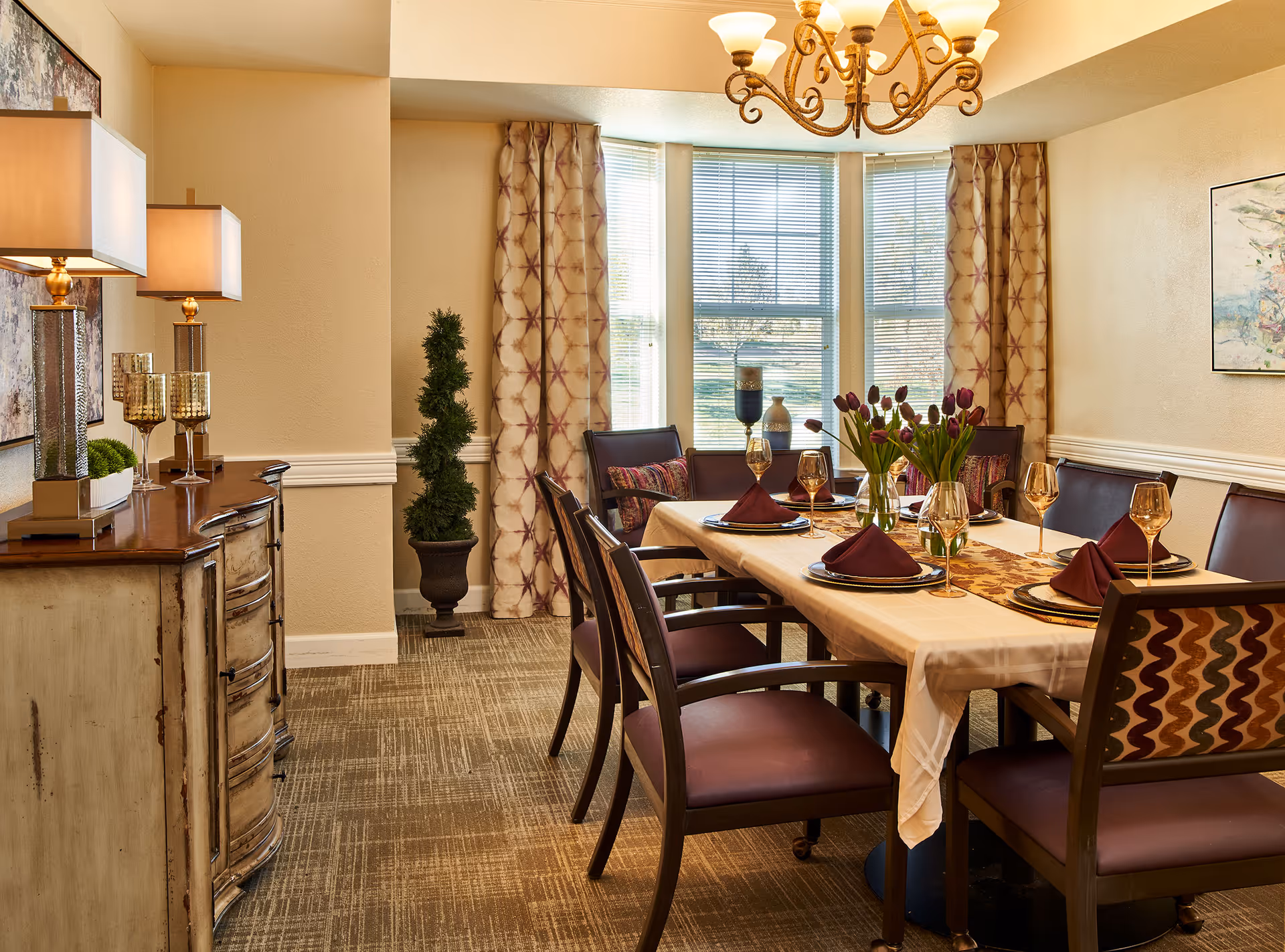 A well-lit dining room with a rectangular table set for six people. The table is covered with a beige tablecloth and decorated with a floral centerpiece and folded burgundy napkins on plates. Surrounding the table are wooden chairs with cushioned seats and backs. The room features large windows with patterned curtains, a chandelier overhead, a sideboard with lamps and decorative items, and a potted plant in the corner.
