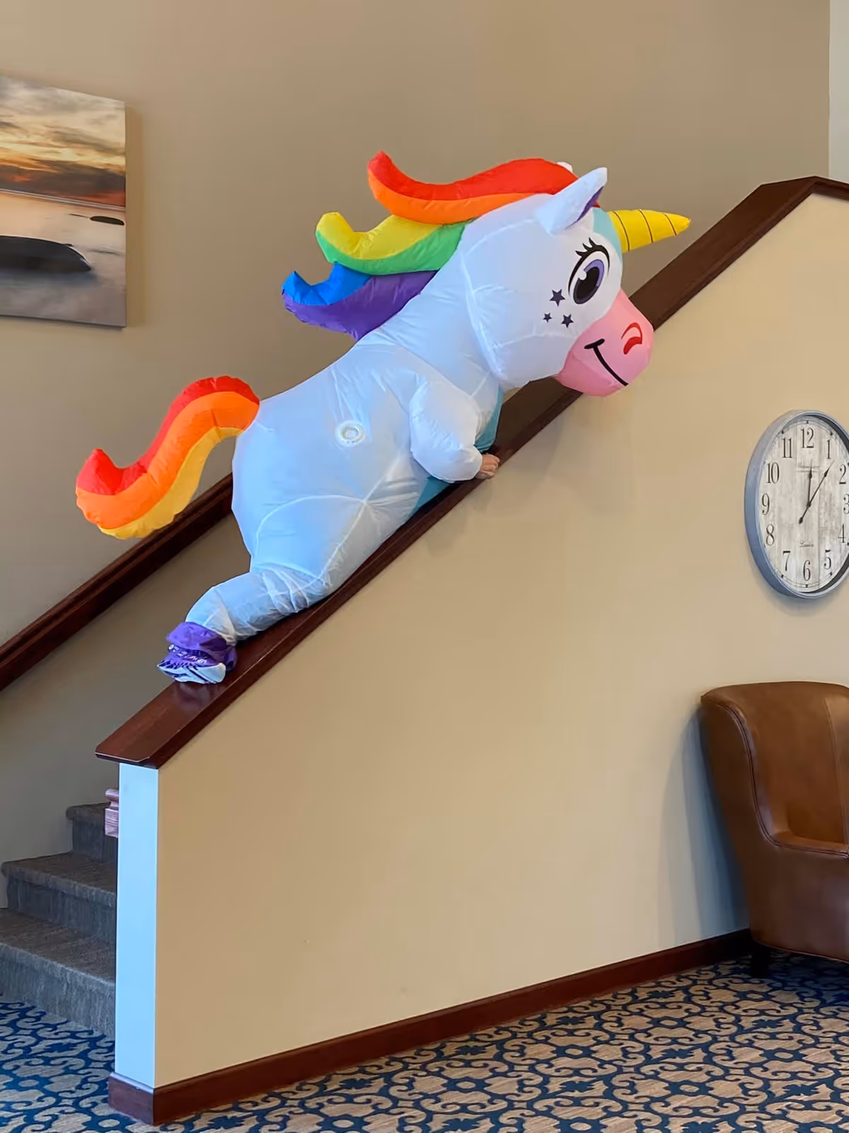 An inflatable unicorn costume with a rainbow mane and tail is worn by a person climbing a carpeted staircase with a wooden handrail. The setting appears to be an interior space with beige walls, a blue patterned carpet, a brown leather chair, a round wall clock showing 12:05, and a landscape painting on the wall.