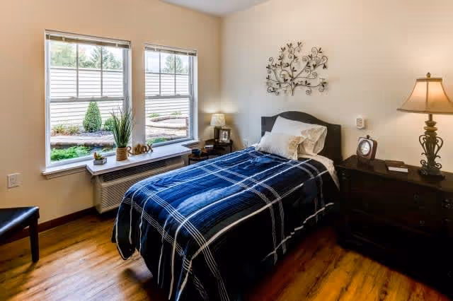 A cozy bedroom with a single bed covered in a blue plaid blanket and white pillows. The room has wooden flooring, two windows with a view of greenery outside, a small table with decorative items including a plant and the word 'Relax', a nightstand with a lamp and clock, and a wall decoration above the bed.