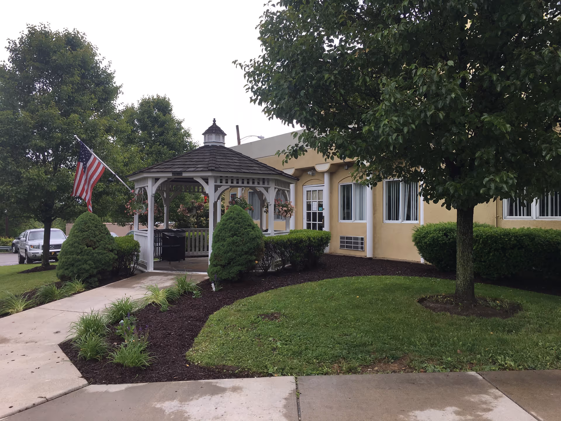 Outdoor view of a senior living facility showing a white gazebo with hanging flower baskets, surrounded by green bushes and trees. An American flag is displayed on a pole near the gazebo. The building in the background has beige walls and several windows with white frames.