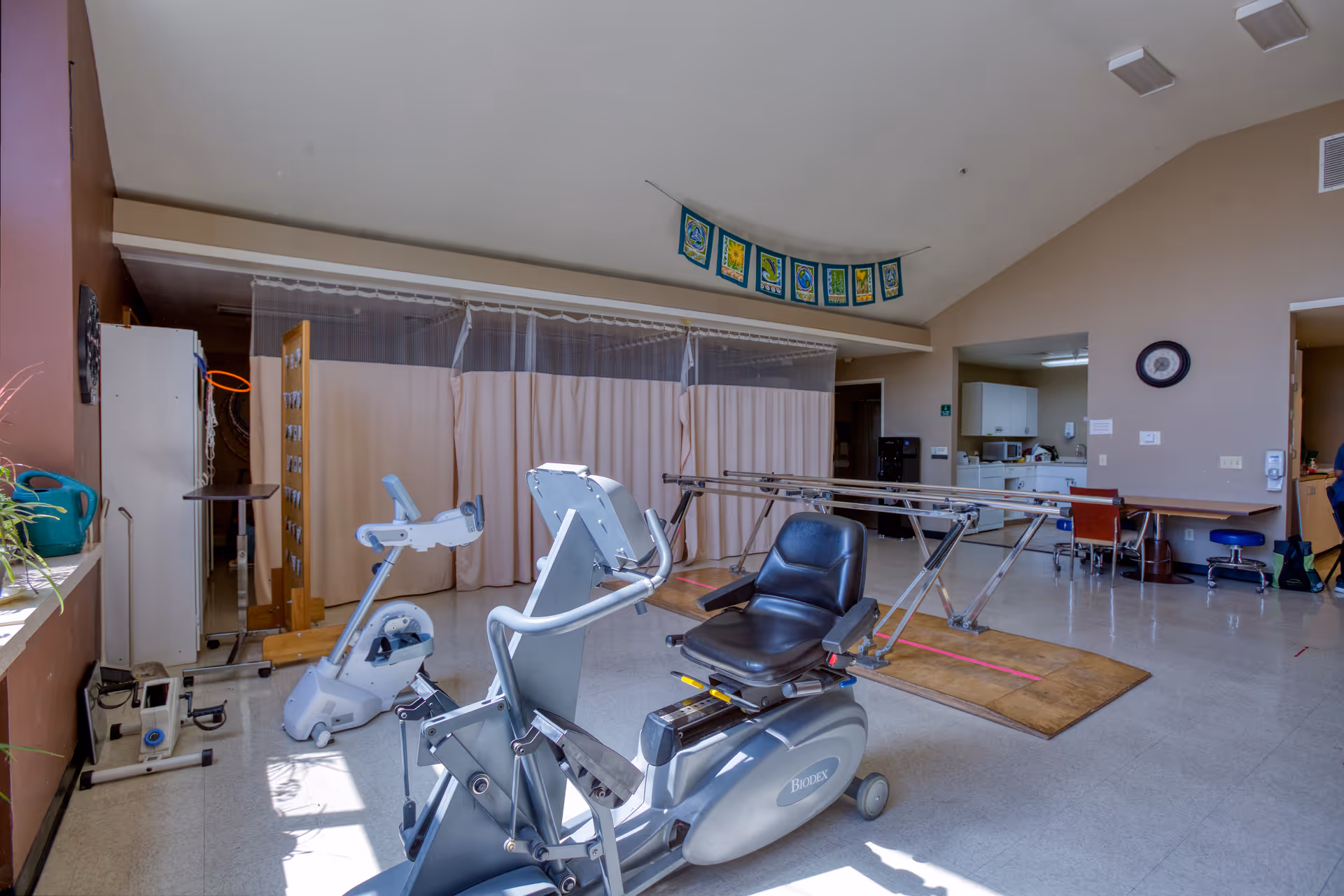 A rehabilitation or physical therapy room with exercise equipment including a recumbent bike and parallel bars. The room has beige walls, a high ceiling, and a curtain divider. There is a desk with chairs and cabinets in the background.