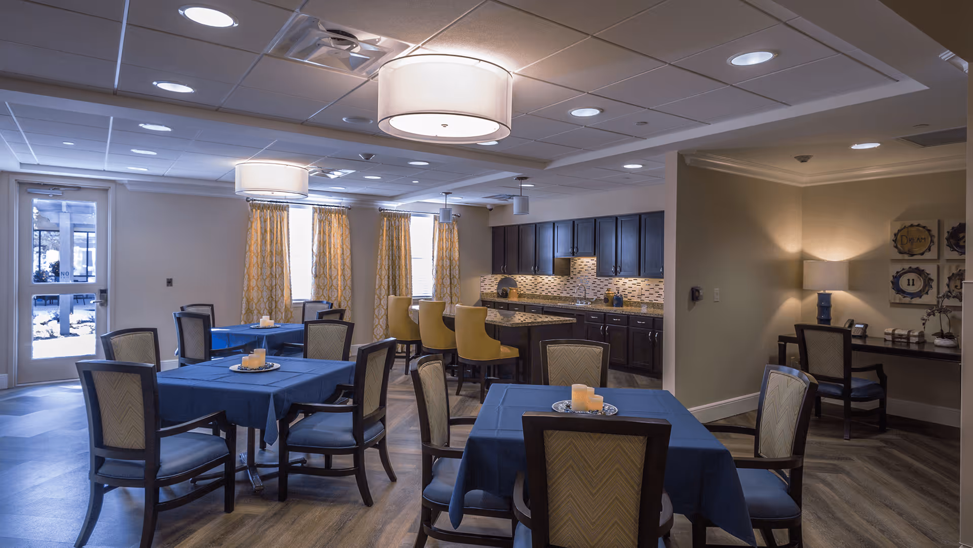 Interior view of a dining area in a senior living facility with several tables covered in blue tablecloths, each with a centerpiece of candles. The room features wooden chairs, large windows with yellow patterned curtains, a kitchen area with dark cabinets and a tiled backsplash, and a small desk with a lamp in a corner.