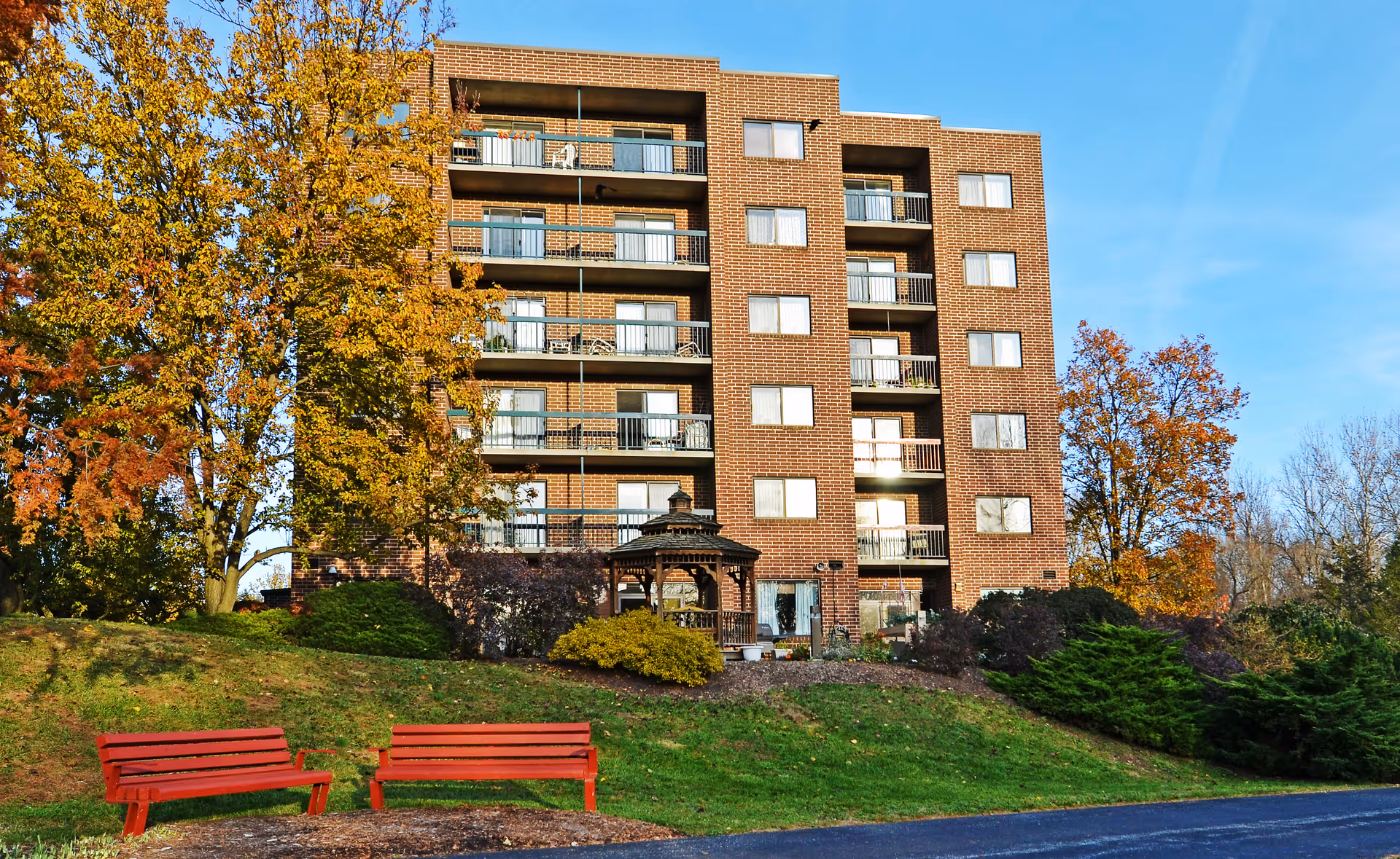 Front view of a multi-story brick senior living building with balconies, a gazebo, red benches, and autumn trees.