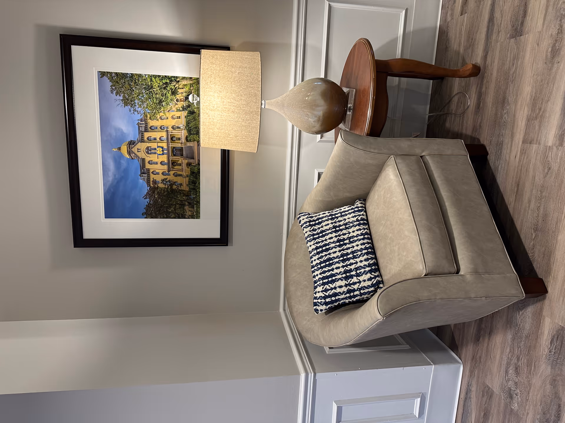 A cozy corner with a beige armchair featuring a blue and white patterned pillow, next to a wooden side table with a beige lamp. Above the chair is a framed photograph of a historic yellow building with a golden dome, surrounded by trees under a blue sky.