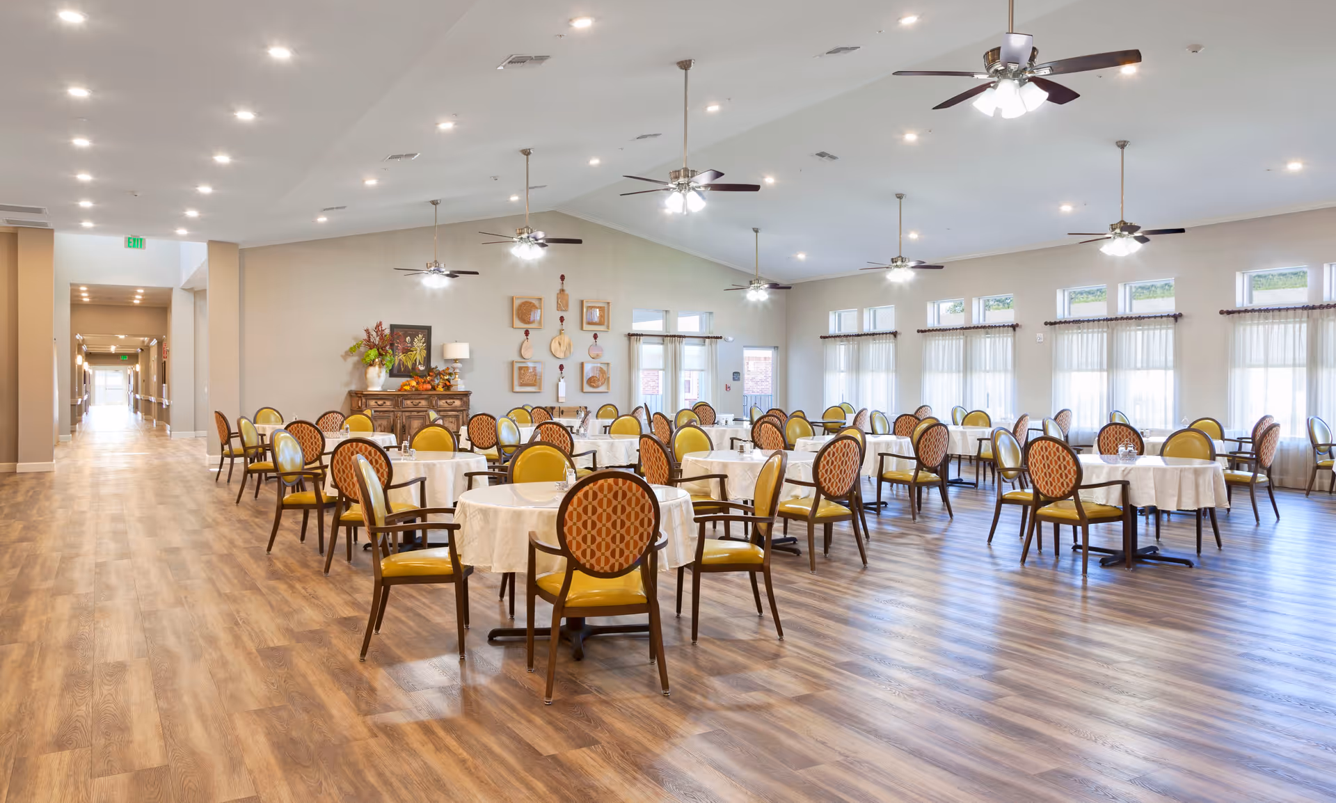 A spacious dining room with multiple round tables covered with white tablecloths, each surrounded by chairs with yellow seats and patterned backs. The room has wood flooring, ceiling fans with lights, large windows with sheer curtains, and a decorative sideboard with floral arrangements and wall art.
