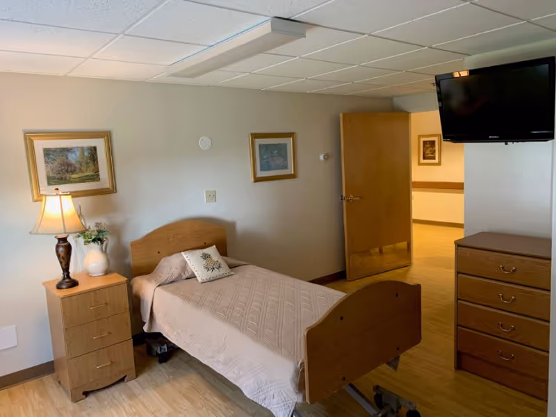 A simple resident bedroom with a single wooden hospital-style bed, nightstand with lamp, dresser, wall-mounted TV, and an open door to the hallway.