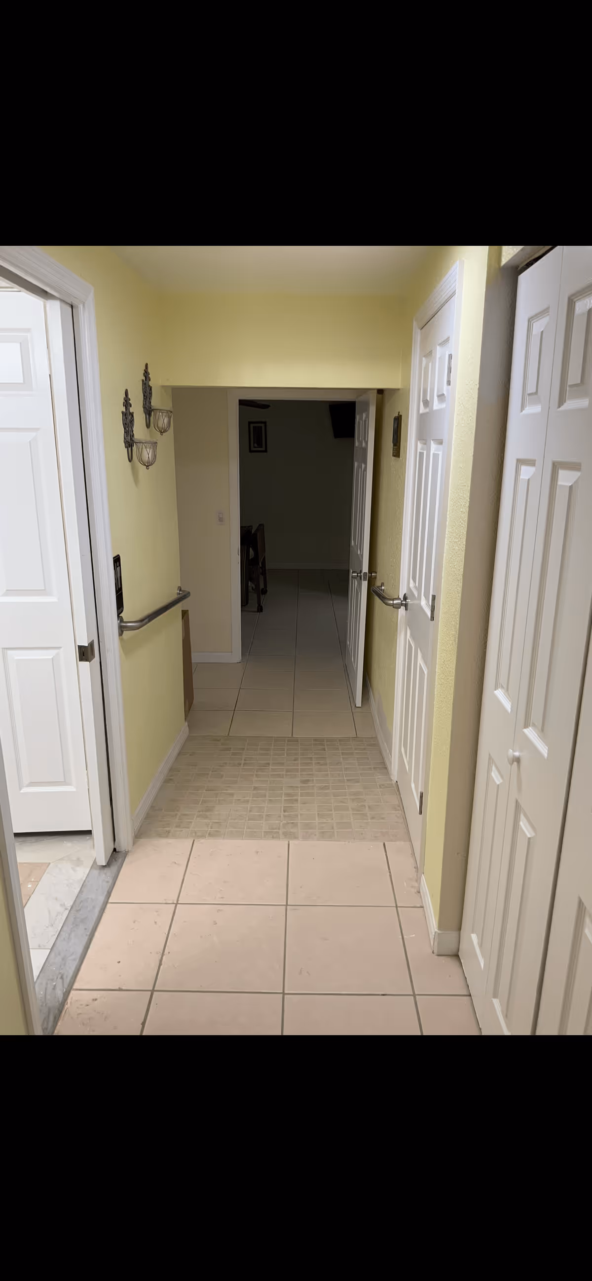 A narrow hallway with light yellow walls and tiled floors. There are white doors on both sides of the hallway, some open and some closed. Metal handrails are mounted on the walls on both sides. The hallway leads to a darker room in the background with some furniture visible.