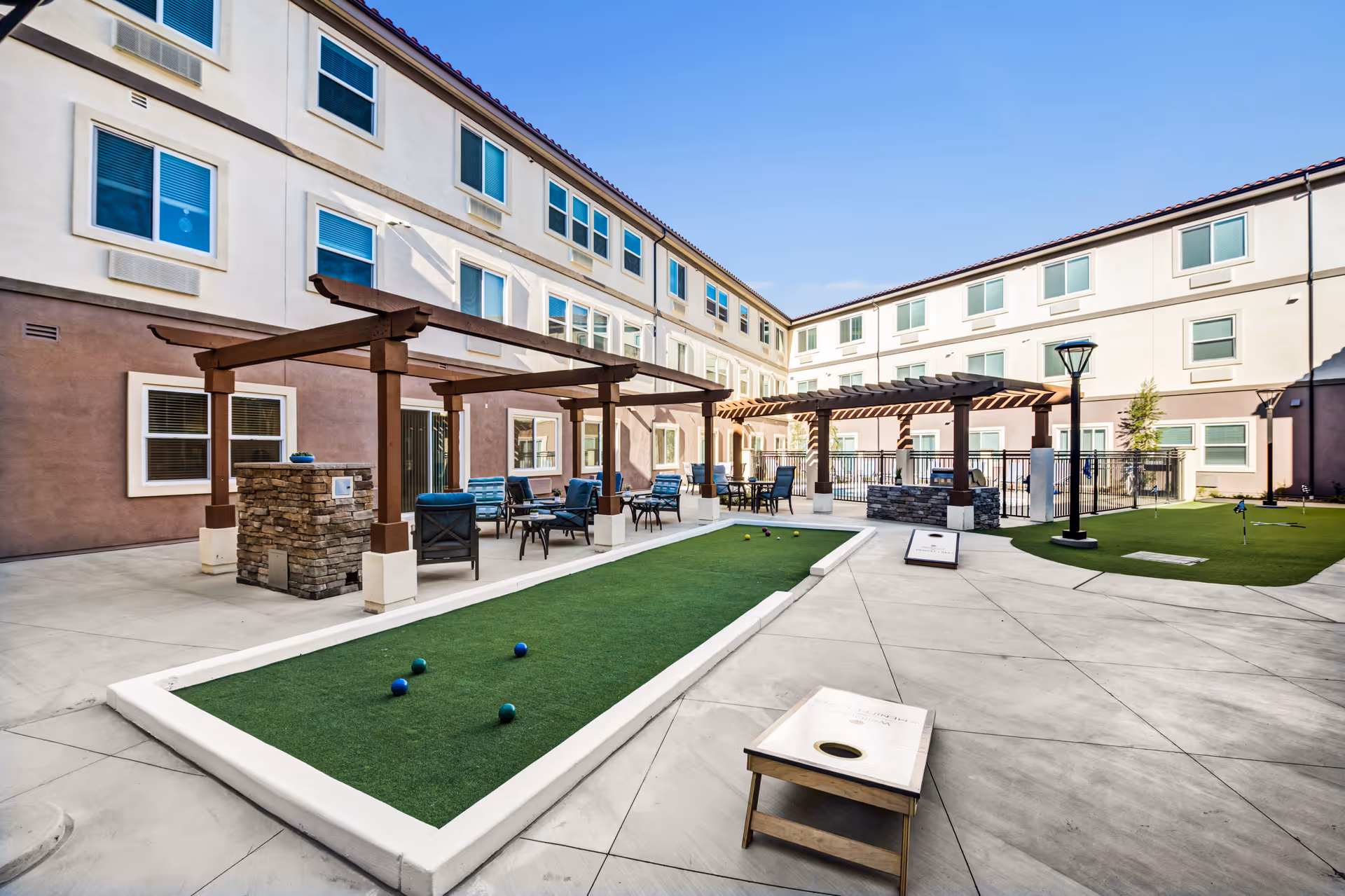 Sunlit courtyard of a senior living facility with pergolas, outdoor seating, bocce/putt lawns and cornhole boards surrounded by a three-story building.