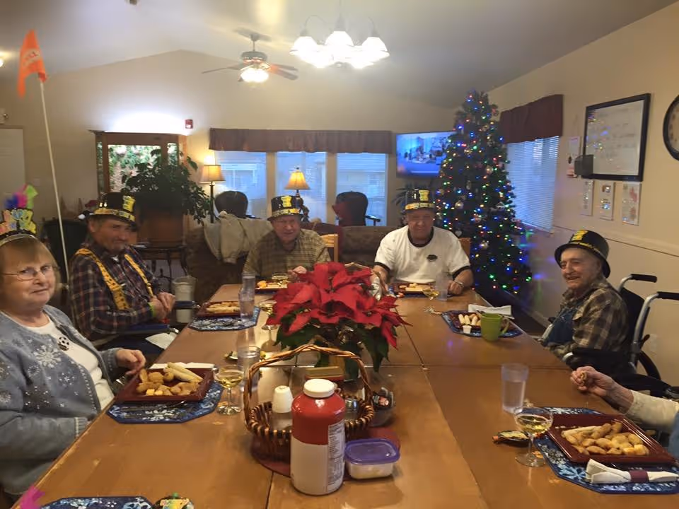 A group of elderly people sitting around a large wooden dining table in a decorated room. They are wearing festive hats and enjoying a meal together. A Christmas tree with colorful lights is visible in the background, along with a television and some windows. A poinsettia plant is placed in the center of the table.