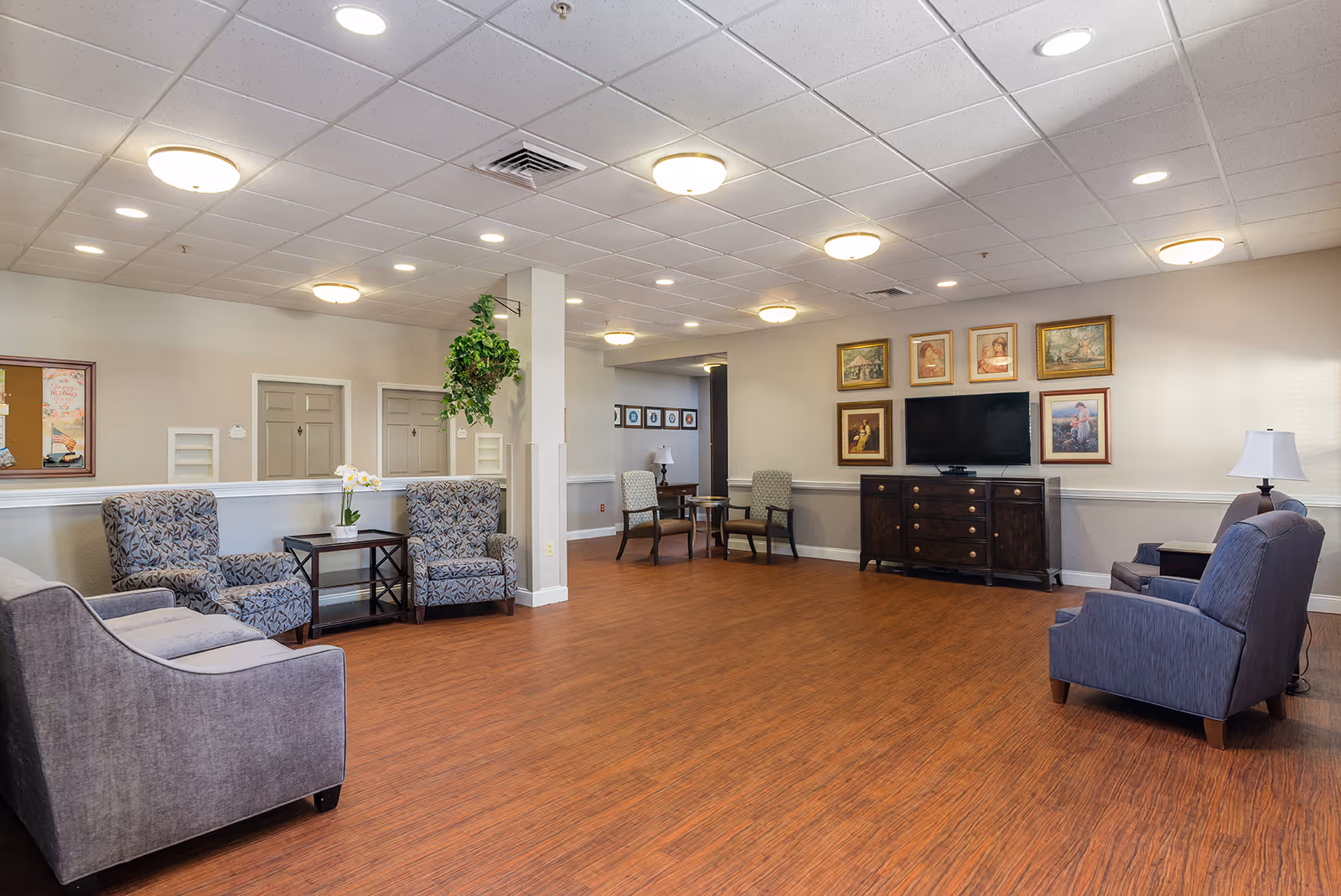 A spacious senior living facility common area with wood flooring, multiple upholstered chairs and sofas arranged around small tables, a flat-screen TV mounted on a dark wooden cabinet, framed artwork on the walls, and ceiling lights providing bright illumination.