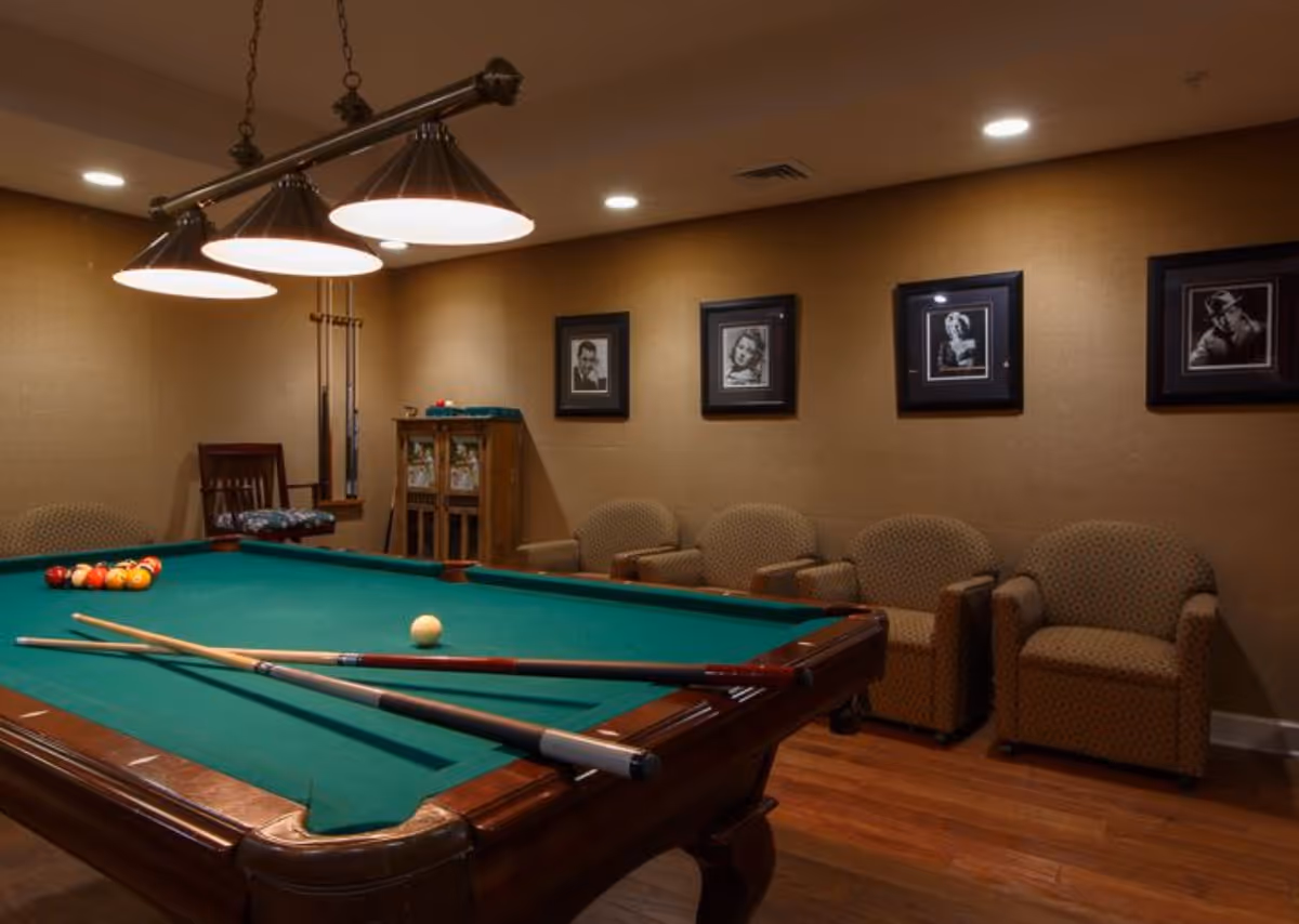 A cozy game room with a green felt pool table in the center, two pool cues resting on the table, and a set of billiard balls arranged for a game. The room has warm lighting from a three-shade hanging lamp above the table. Along one wall, there are four patterned armchairs and framed black-and-white portraits hanging above them. The floor is wooden, and there is a small cabinet and a chair in the corner.
