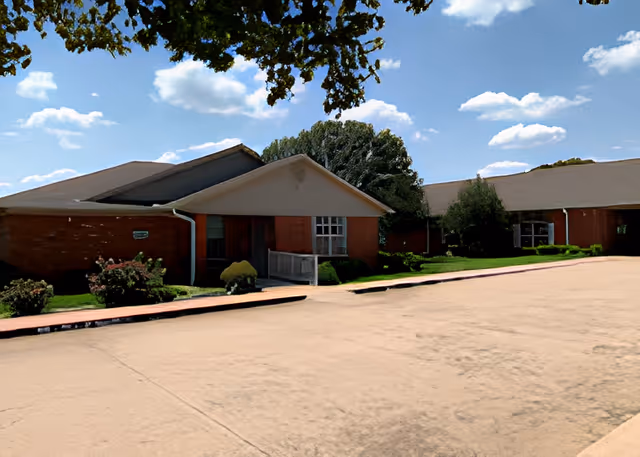 Exterior view of a single-story brick building with a sloped roof, surrounded by green shrubs and trees under a blue sky with scattered clouds.