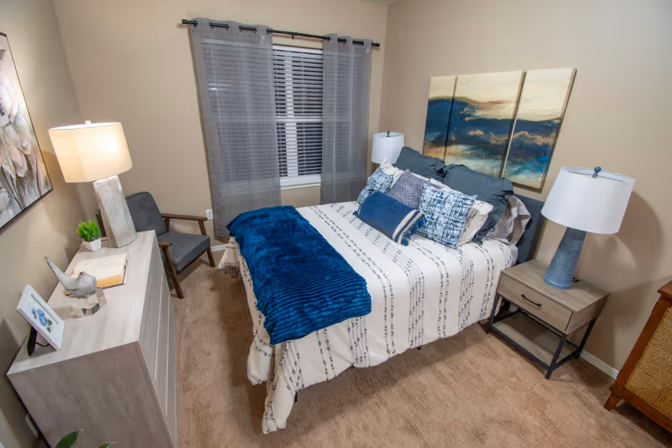 A cozy bedroom with a bed dressed in white and blue patterned bedding and multiple pillows. A blue throw blanket is folded at the foot of the bed. On the right side of the bed is a nightstand with a blue lamp. On the left side of the room is a dresser with a lamp, a small plant, a decorative bird, and a framed picture. A gray chair is positioned near the window, which has gray curtains. Above the bed is a three-panel abstract painting with blue and beige tones. The room has beige walls and carpeted floor.