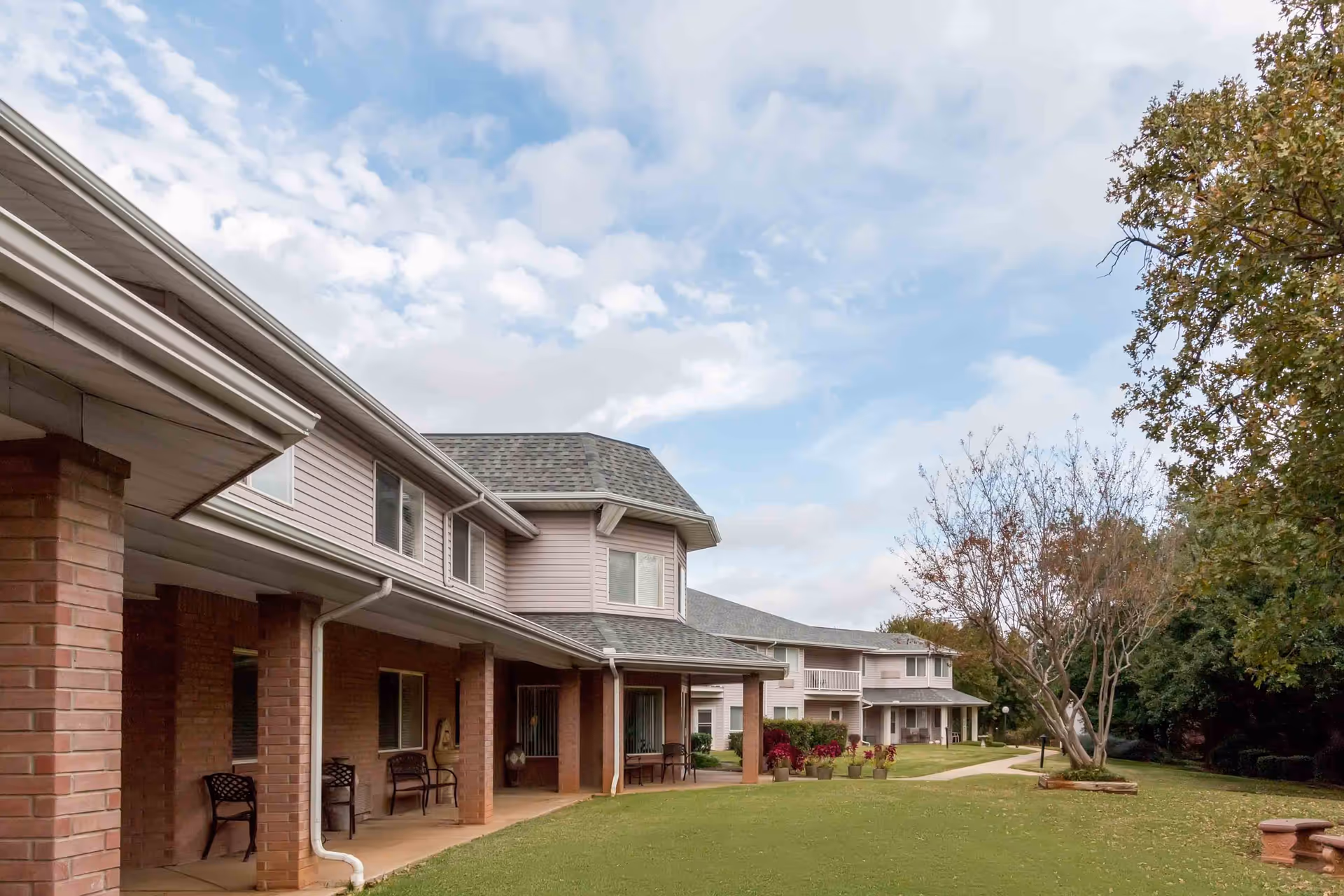 Two-story senior living building with a covered porch, patio chairs, a walkway and a grassy lawn with trees.