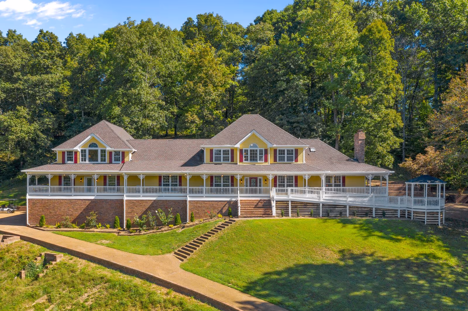 A large two-story yellow building with red shutters and a wide white porch railing, surrounded by lush green trees and a well-maintained lawn, under a clear blue sky.