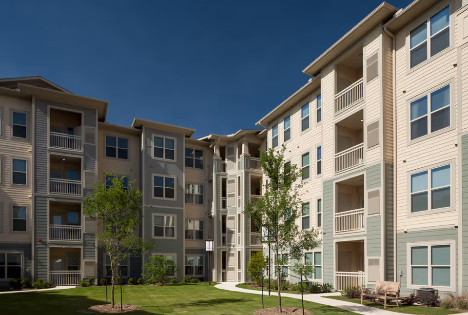 Courtyard and facade of a multi-story senior living apartment building with balconies, small trees, lawn, and a bench.