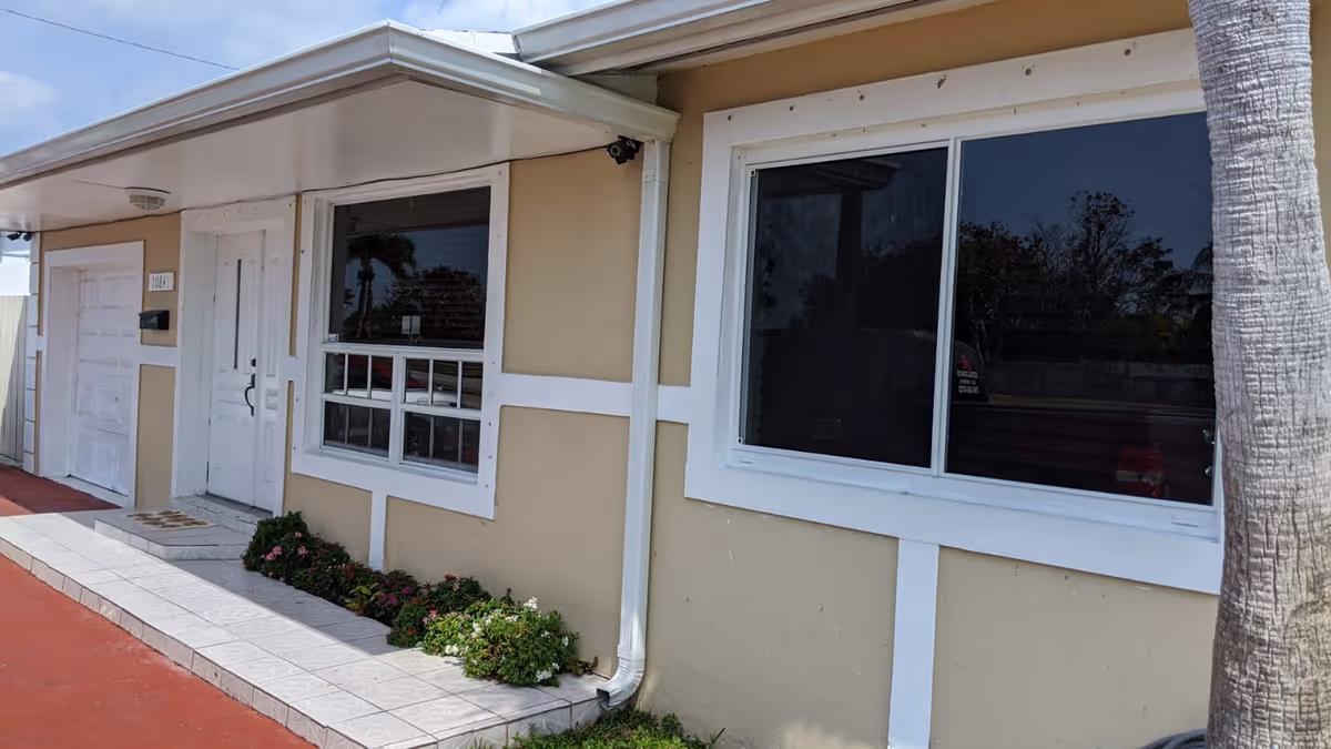 Exterior view of a single-story building with beige walls and white trim. There is a white door with a small window next to it, a garage door, two windows, and a small garden bed with flowers along the tiled walkway. A palm tree is visible on the right side of the image.