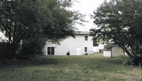View of the backyard of a two-story white house with a grassy lawn and trees on both sides. There is a small shed on the right side near the house.