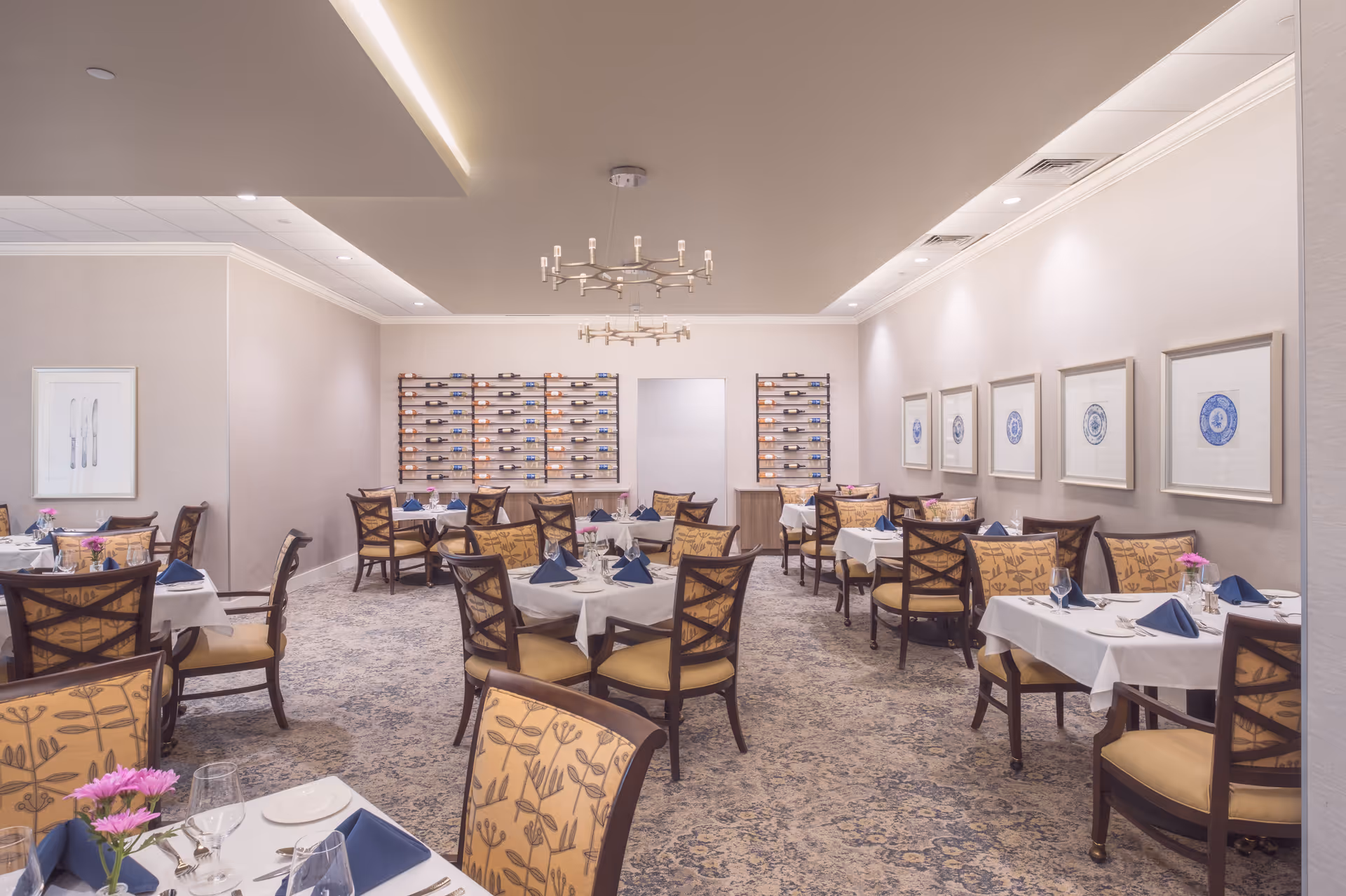 A well-lit dining room with multiple tables covered in white tablecloths, each set with plates, glasses, silverware, and blue folded napkins. The chairs have patterned upholstery, and there are framed artworks on the walls. Two wine racks filled with bottles are mounted on the far wall, and a modern chandelier hangs from the ceiling.