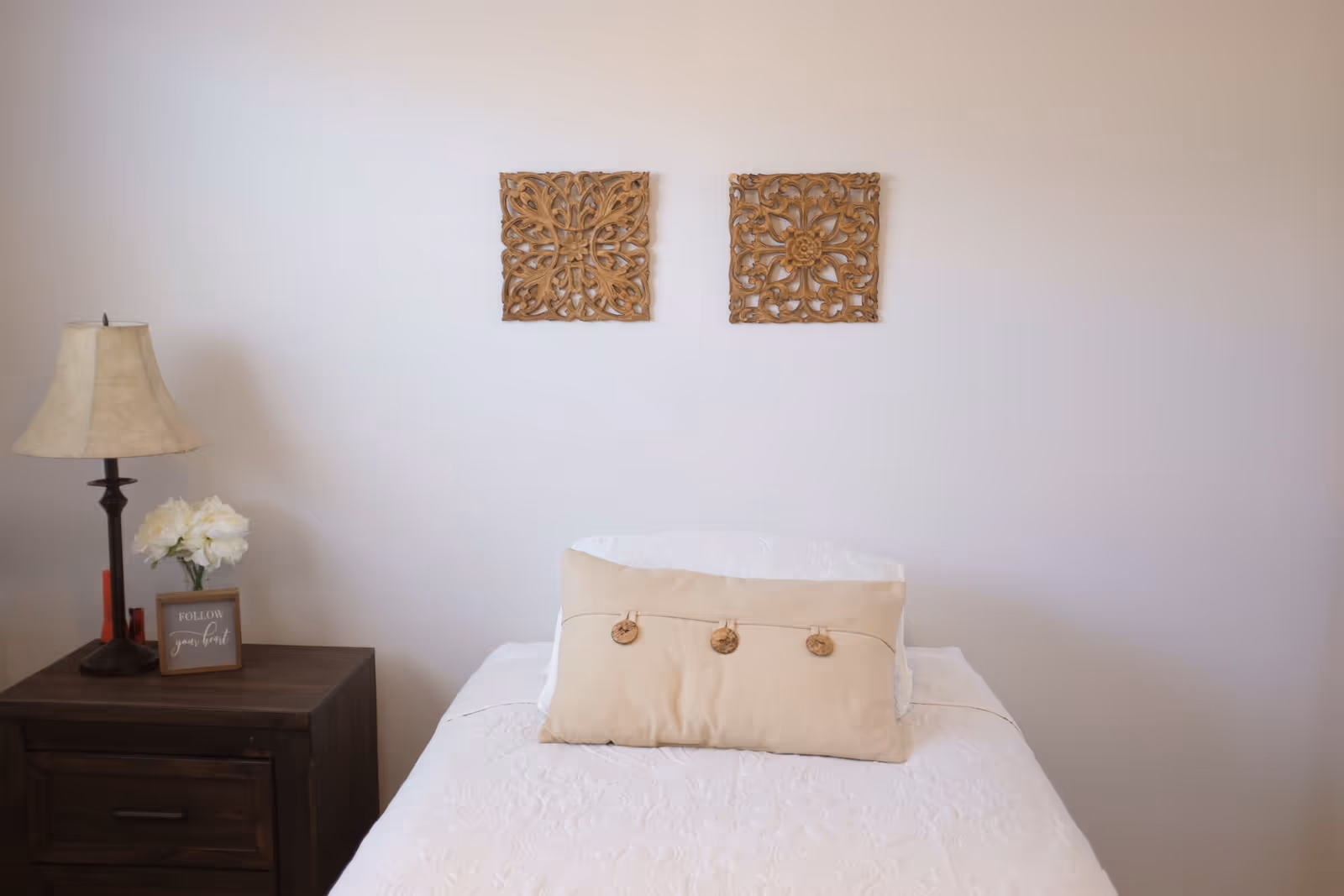 A neatly made single bed with a beige decorative pillow beneath two carved wooden wall hangings and a bedside table with a lamp and flowers.