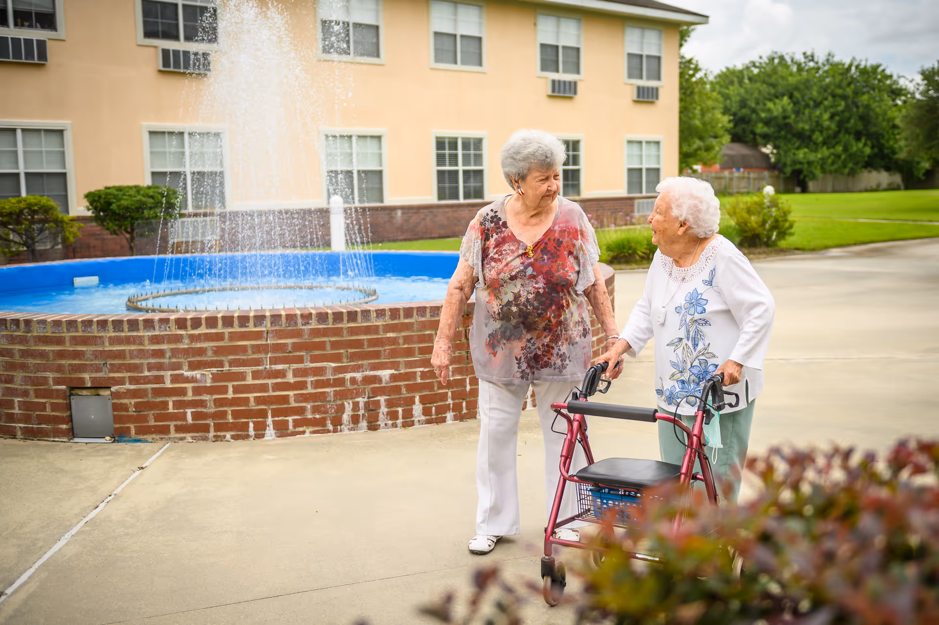 Two elderly women walking and talking outside near a brick fountain with water spraying up. One woman is using a walker. They are in front of a building with multiple windows and surrounded by greenery.