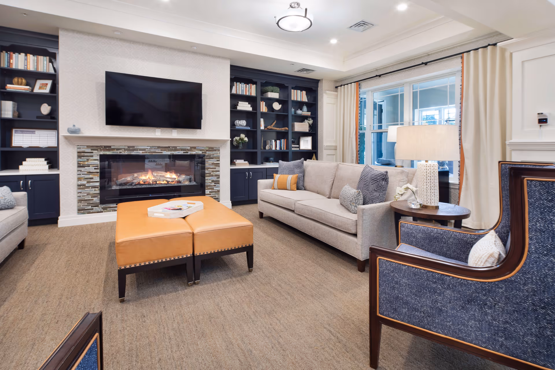 A cozy living room with a beige sofa adorned with decorative pillows, two blue armchairs with wooden frames, and a pair of tan ottomans in the center. A modern fireplace with a tiled surround is below a mounted flat-screen TV. Built-in dark blue bookshelves flank the fireplace, filled with books and decorative items. Large windows with cream-colored curtains allow natural light to fill the room.