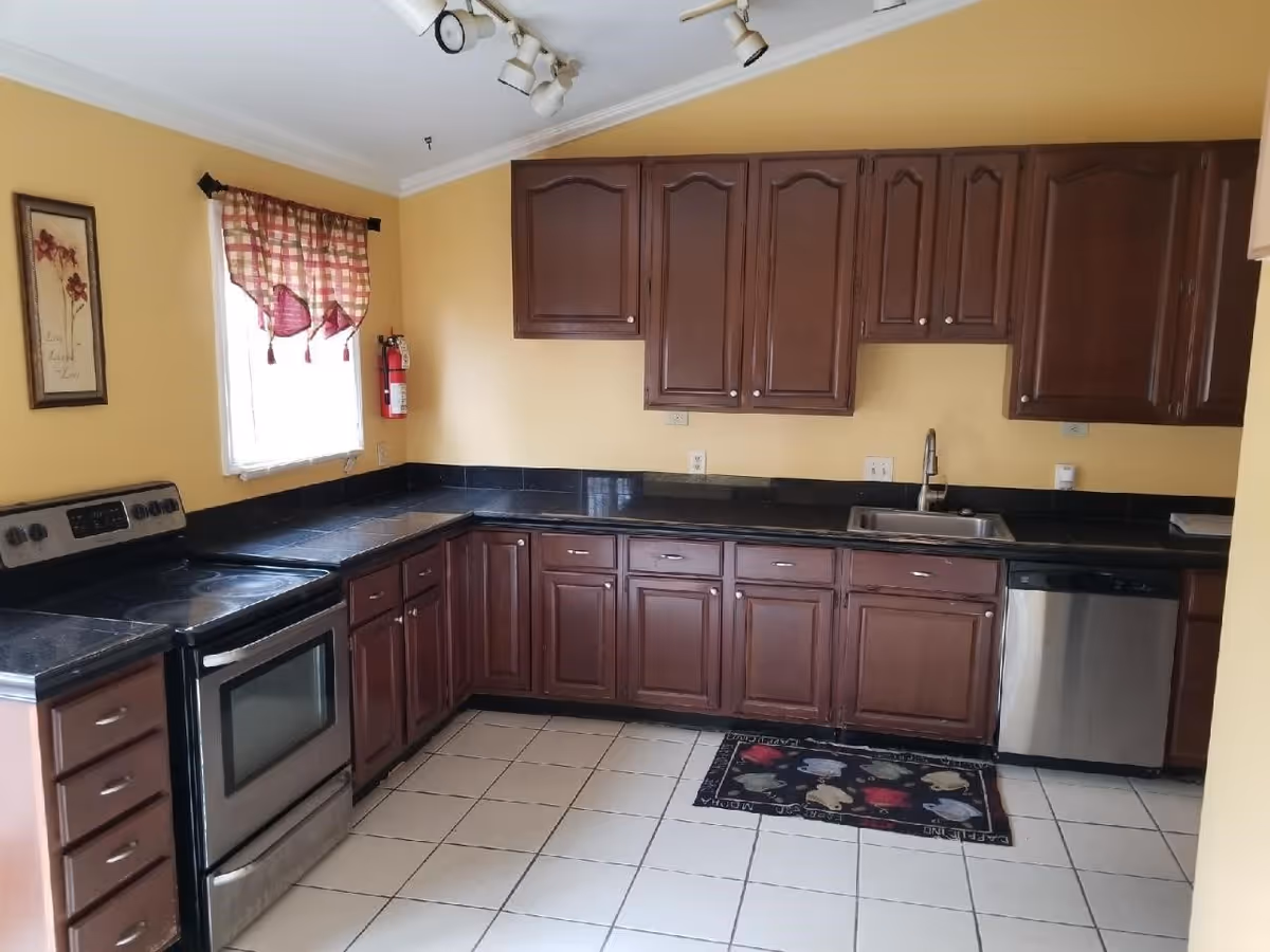 A kitchen with dark wooden cabinets, black countertops, a stainless steel stove, dishwasher, and a sink. The walls are painted yellow, and there is a window with a red and white checkered curtain. A fire extinguisher is mounted on the wall near the window, and a decorative rug is placed on the tiled floor.
