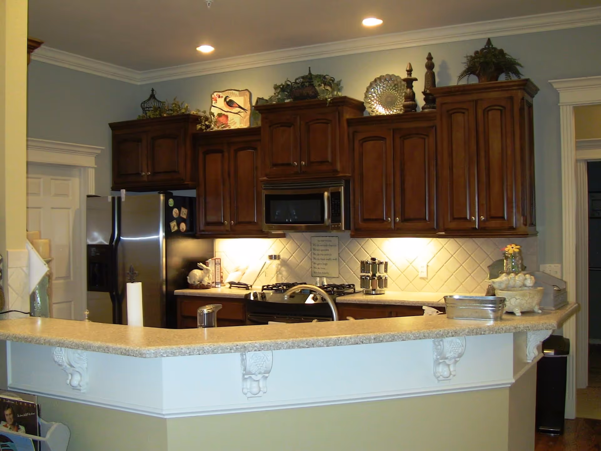 A kitchen area with dark wooden cabinets, a stainless steel refrigerator, a microwave above a stove, and a beige countertop with decorative corbels. The backsplash is tiled in a diamond pattern, and there are various decorative items on top of the cabinets and countertop. The kitchen is well-lit with recessed ceiling lights and under-cabinet lighting.