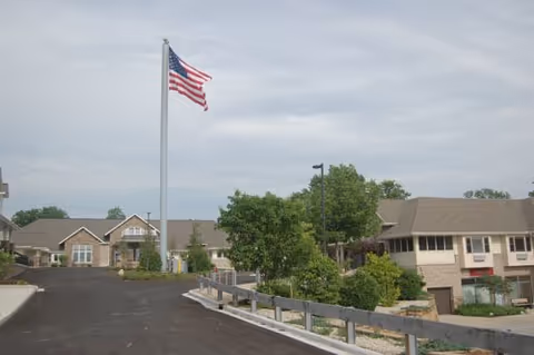 Driveway leading to the front of a senior living complex with an American flag on a tall flagpole and low brick buildings.