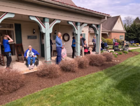 Residents and staff gathered on a covered front porch of a senior living facility with landscaping and a lawn in front.