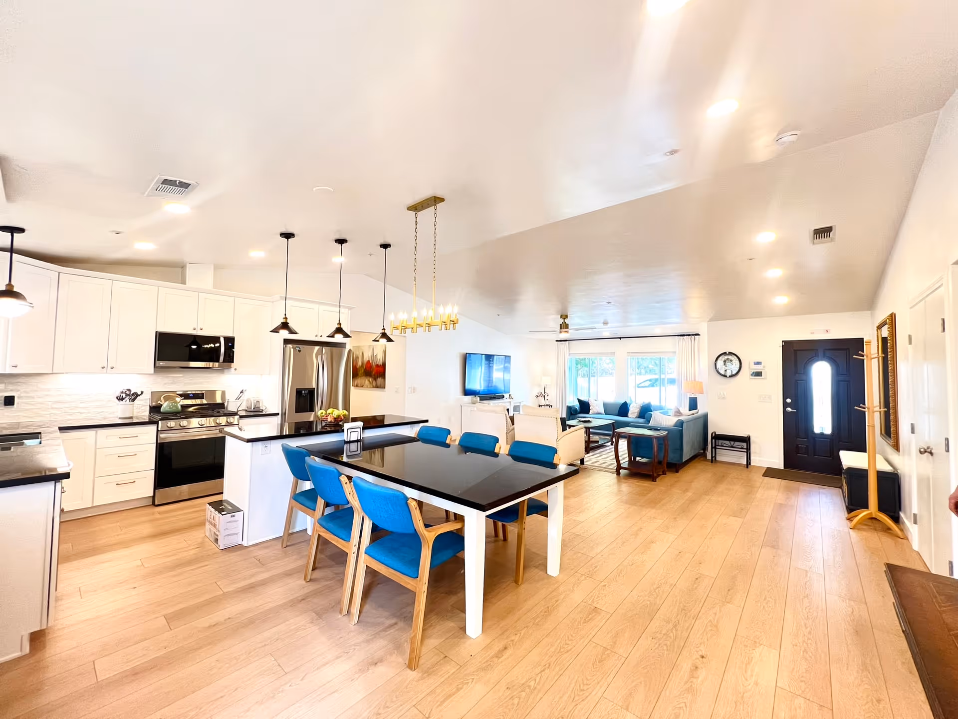 Bright and spacious open-concept interior of a senior living facility showing a modern kitchen with white cabinets, stainless steel appliances, and a black countertop island. Adjacent to the kitchen is a dining area with a black table and blue cushioned chairs. In the background, there is a living room area with a blue sectional sofa, a wall-mounted TV, and large windows letting in natural light. The space features light wood flooring and white walls, with a black front door visible on the right side.