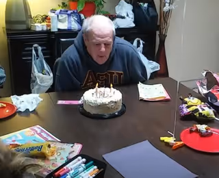 An elderly man wearing a dark hoodie is seated at a table, blowing out candles on a birthday cake. The table has various items including markers, papers, and snacks. The background shows bags and other items on a counter and a vase with decorative branches.