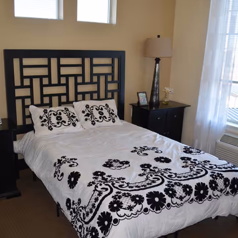 Neatly made bed with a black geometric headboard and white duvet with black floral pattern in a bright bedroom with nightstands and a lamp.