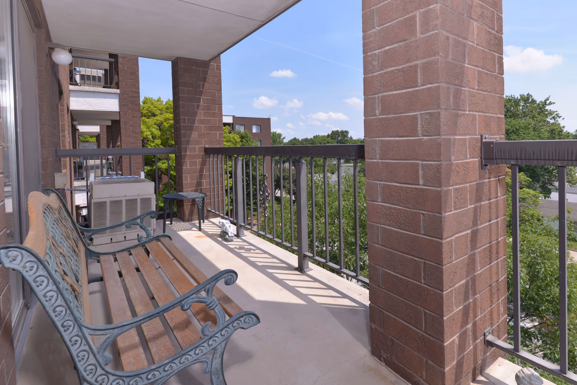 A balcony with a decorative metal and wooden bench, a small black metal table, and an air conditioning unit. The balcony has brick pillars and a metal railing, overlooking green trees and a clear blue sky with some clouds.