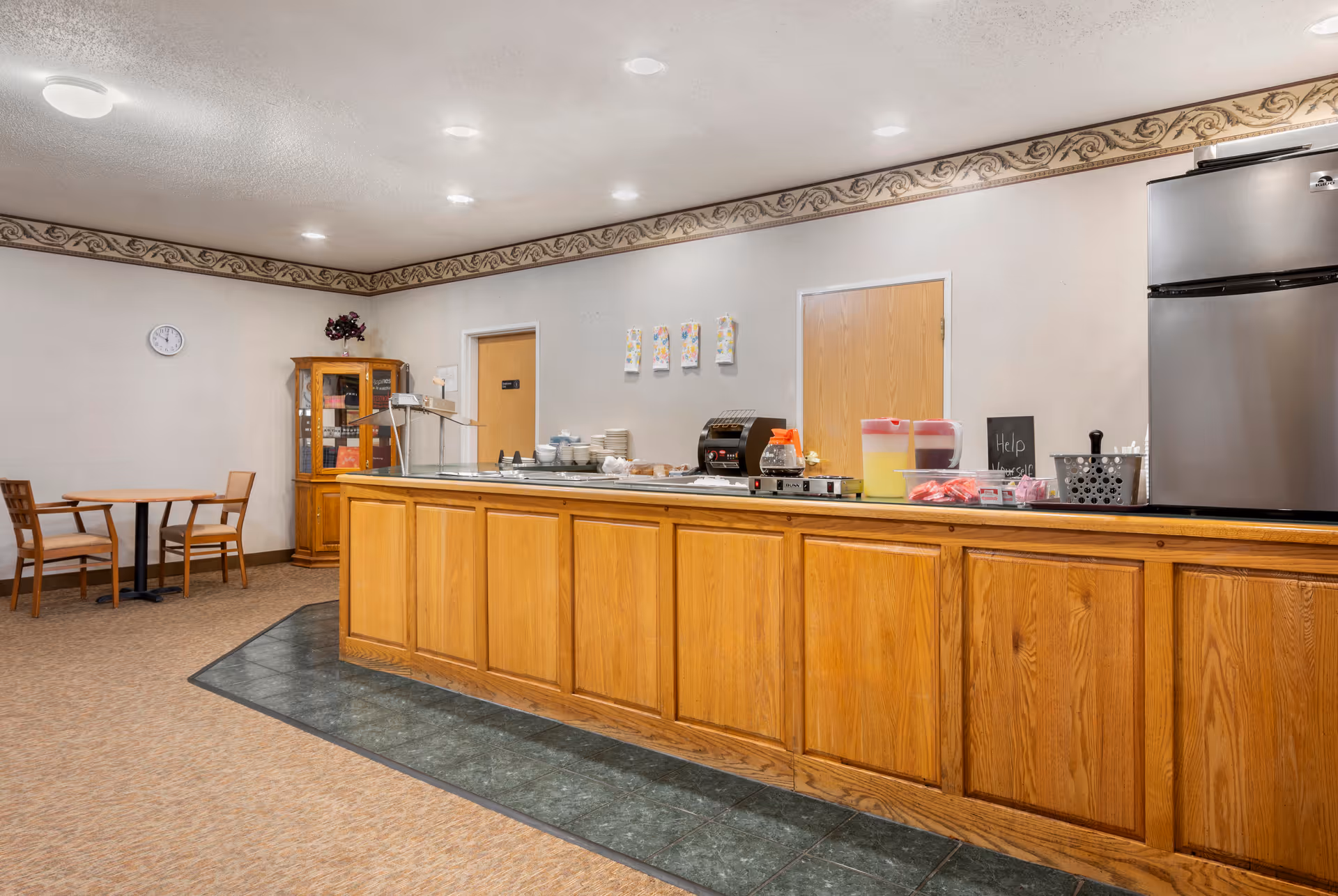 Communal dining area with a long wooden serving counter, beverage dispensers, small tables and chairs, and a refrigerator.