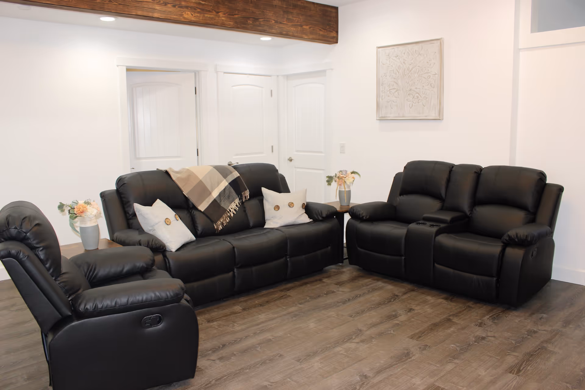 A cozy living room area with two black leather sofas and one black leather recliner. The sofas have light-colored pillows and a plaid throw blanket. There are two small side tables with flower vases on each side of the sofas. The room has white walls, wooden flooring, and three white doors in the background. A decorative wall art piece hangs on the wall above one sofa.