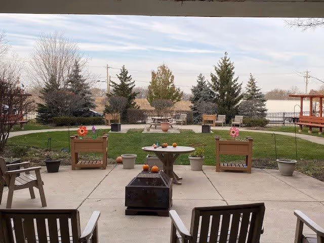 Outdoor patio area with wooden chairs, small tables, potted plants, and decorative flowers. The patio overlooks a grassy area with trees and shrubs, under a cloudy sky.