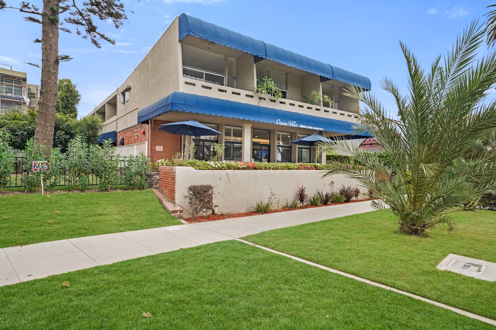 Two-story senior living building with blue awnings, patio umbrellas, and a landscaped front lawn.