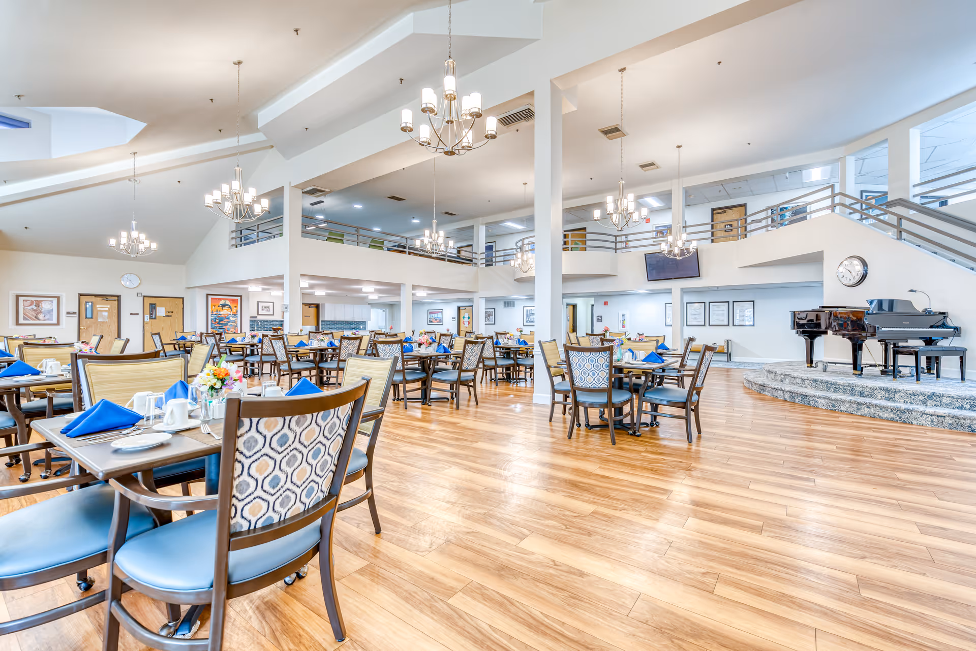 Spacious dining room with set tables and chairs, chandeliers, a raised stage with a piano, and an upper mezzanine.